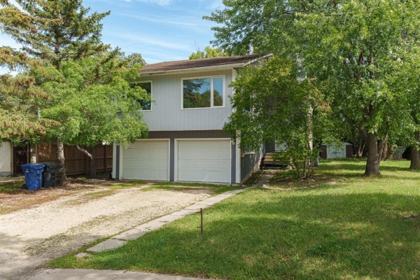 Side view of the light blue house showing the double garage with two white doors and two cars on the gravel driveway. - 7 Scotswood Drive, Winnipeg
