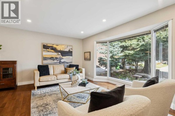 Living room with a bay window overlooking trees, a leather sofa, two armchairs, and a marble coffee table. - 7 Woodgreen Crescent Sw, Calgary