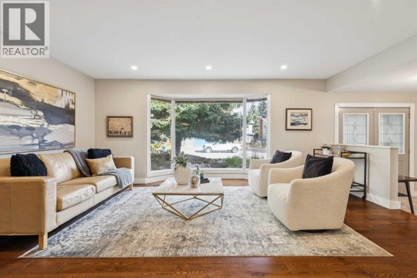 Spacious living room featuring a bay window, large rug, leather sofa, two bouclé armchairs, and hardwood floors. - 7 Woodgreen Crescent Sw, Calgary