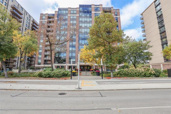 Front exterior of a brick apartment building with lush landscaping and trees. - 703 141 Wellington Crescent, Winnipeg