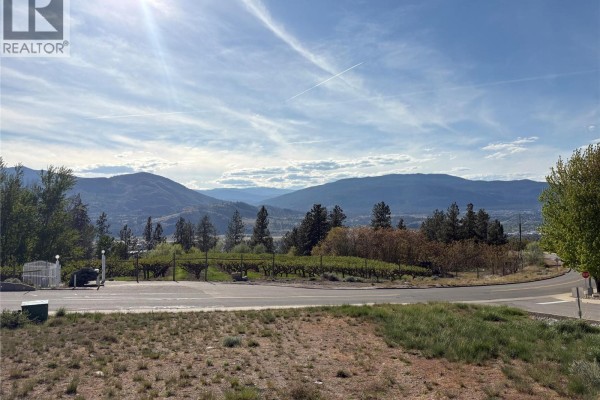 View of a rural landscape with mountains and a lake, showing a road, vineyards, and trees. - 703 Pineview Road, Penticton