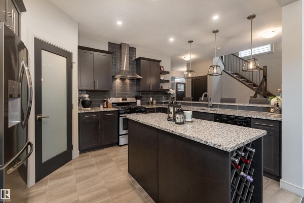 Kitchen with dark cabinetry, island with wine rack, stainless steel refrigerator, and view of staircase. - 7038 172a Av Nw, Edmonton