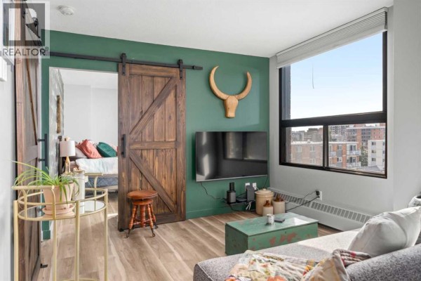 Living room with large windows overlooking the city, a gray sofa, and a rustic wooden barn door. - 704, 1202 13 Avenue Sw, Calgary