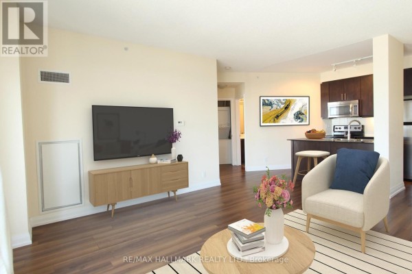Living room with a television, sofa, armchair, coffee table, and kitchen in the background. - 704 - 85 The Donway W, Toronto