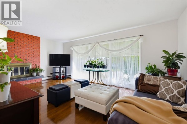 Living room with a brick fireplace, large window with sheer curtains, and hardwood floors. - 704 75 Avenue Sw, Calgary