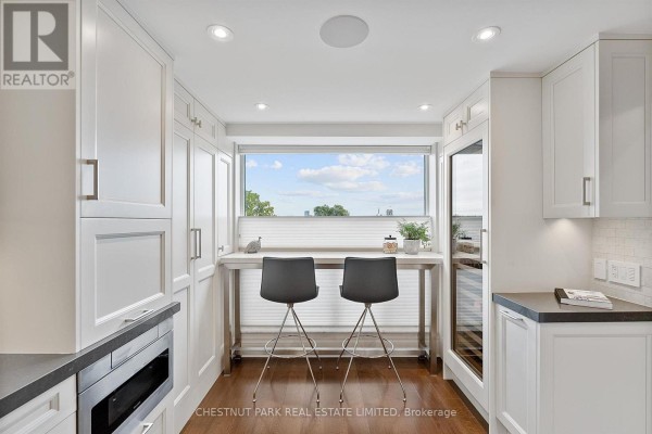 White kitchen cabinets flank a breakfast bar with two stools beneath a wide window with a view. - 706 - 1818 Bayview Avenue, Toronto