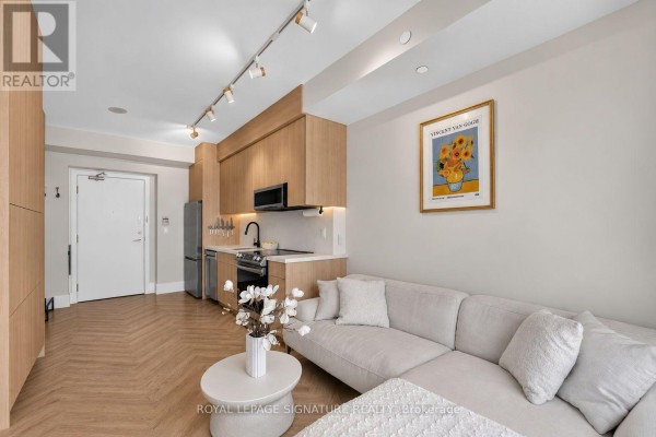Living room adjacent to kitchen featuring a sectional sofa, coffee table, and artwork. - 706 - 33 Shore Breeze Drive, Toronto