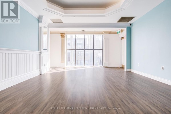 An empty room with light wood laminate flooring, pale blue walls, and large windows overlooking a street. - 706 Pape Avenue, Toronto