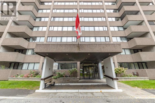 Entrance to the apartment building with a covered overhang, bench seating, and glass doors. - 707 - 3360 Southgate Road, Ottawa