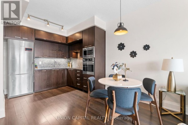 Dining area with a round table and blue chairs, adjacent to a modern kitchen with dark wood cabinets and stainless steel appliances. - 708 - 120 Harrison Garden Boulevard, Toronto