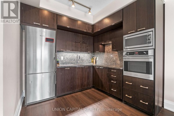 Corner view of a galley kitchen with dark wood cabinets, granite countertops, stainless steel refrigerator, oven, and microwave. - 708 - 120 Harrison Garden Boulevard, Toronto