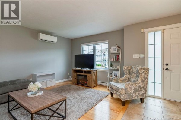 Living room with a grey sectional sofa, floral armchair, electric fireplace, large television, and wood flooring. - 71 Houlahan Street, Dieppe