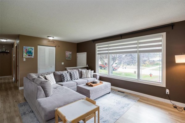 Spacious living room with a grey sectional sofa, large window overlooking the yard, and wood-look flooring. - 71 Rougeau Avenue, Winnipeg