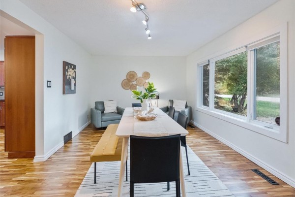 Dining area with a light wood table, bench, chairs, and a large window overlooking the backyard trees. - 713 Buckingham Road, Winnipeg