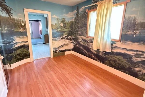 Room with wood-look laminate flooring, scenic mural wallpaper, and a doorway to another room. - 713 Healy Avenue, Radville