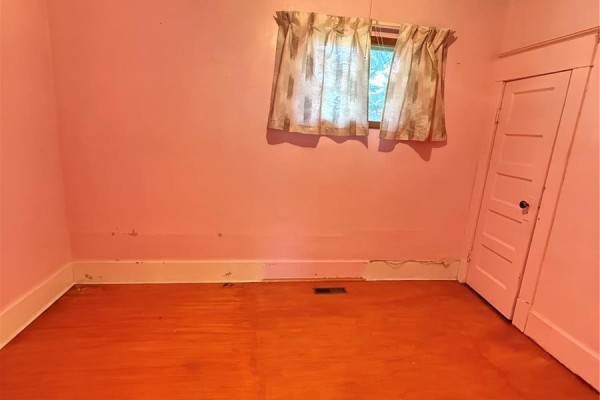 Bedroom with orange-painted wood floor, pink walls, and a window with patterned curtains. - 713 Healy Avenue, Radville