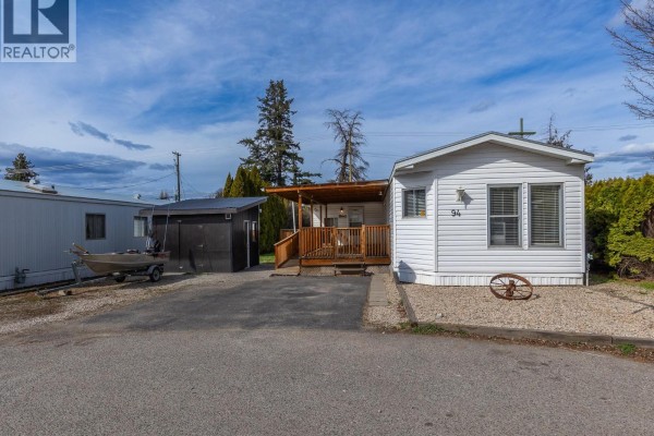 Side exterior view of a white mobile home with a covered wooden deck and a dark shed. - 715 Beaver Lake Road Unit# 94, Kelowna