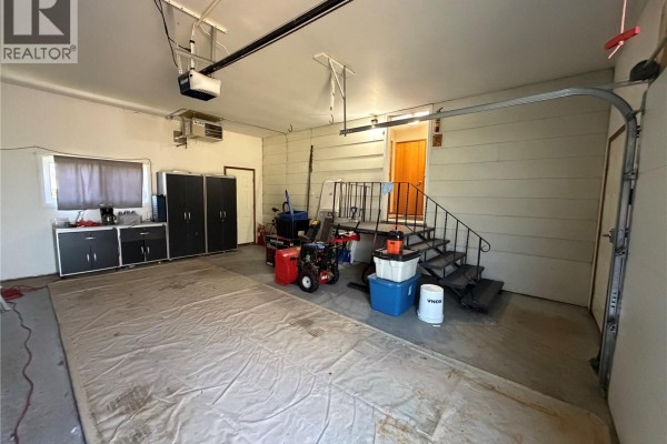 Interior view of a garage showing storage units, a snowblower, a staircase, and a garage door opener. - 715 Mcnaughton Street, Moosomin