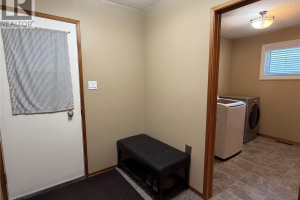 Interior view of a mudroom with a bench, shoe rack, door, and access to a laundry room with washer and dryer. - 715 Mcnaughton Street, Moosomin