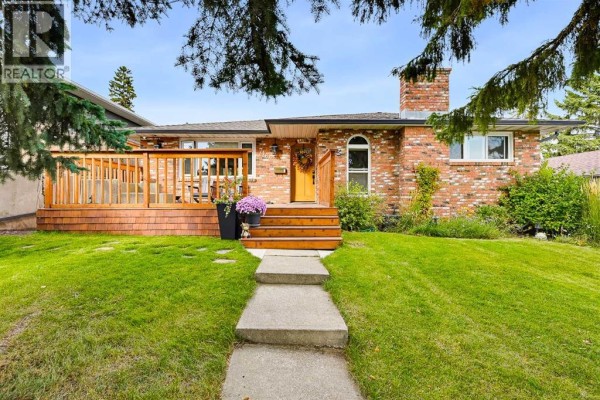 Front view of a brick house with a wooden deck and steps, surrounded by green grass and trees. - 716 Seymour Avenue Sw, Calgary