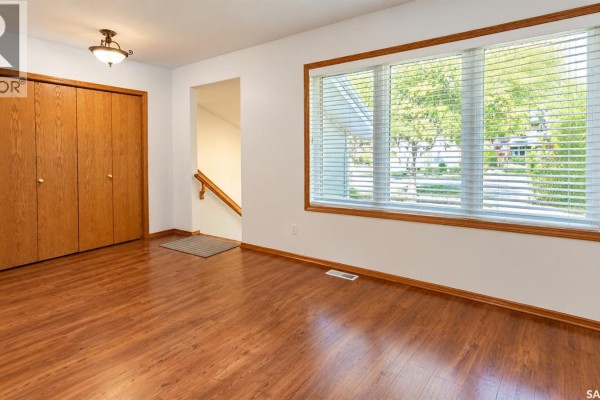 Empty room with wood laminate flooring, a large window with blinds, and wooden closet doors. - 717 31st Street W, Saskatoon