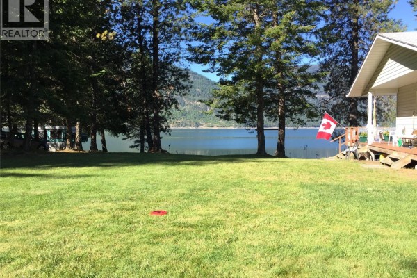 Grassy yard facing a lake with a small building, deck, and Canadian flag visible. - 7180 Little Cayuse Creek Road, Castlegar