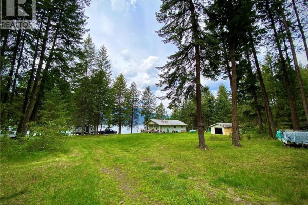 Wide shot of the property showing the house, outbuildings, vehicles, and lake through trees. - 7180 Little Cayuse Creek Road, Castlegar