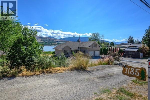View of the house from the driveway, showing the property and a distant lake view. - 7185 Old Kamloops Road, Vernon