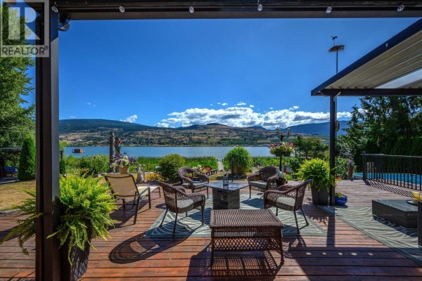 Covered outdoor patio with wicker furniture, a fire pit, and a deck overlooking a lake and hills. - 7185 Old Kamloops Road, Vernon