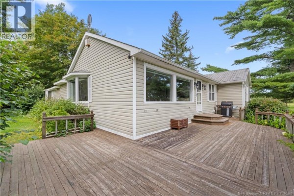 Rear exterior of a light beige house with a large wooden deck and surrounding mature trees. - 72 Hanington, Shediac Cape