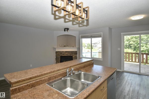 Kitchen island with double sink overlooking a living room with a stone fireplace and patio doors. - 72 Hartwick Ld, Spruce Grove