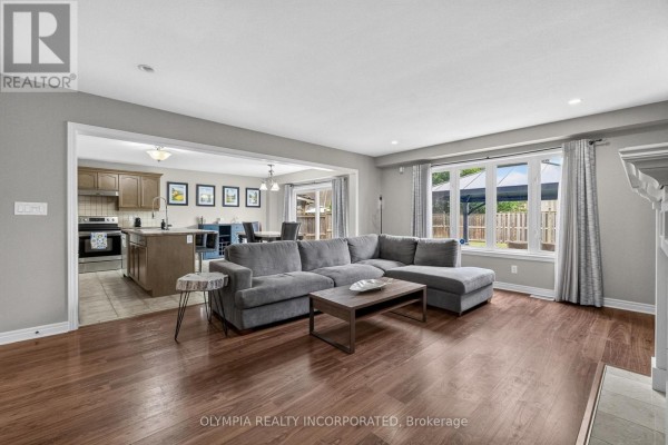 Living room with view into kitchen, gray sectional sofa, hardwood floors, and large window. - 72 Tuliptree Road, Thorold