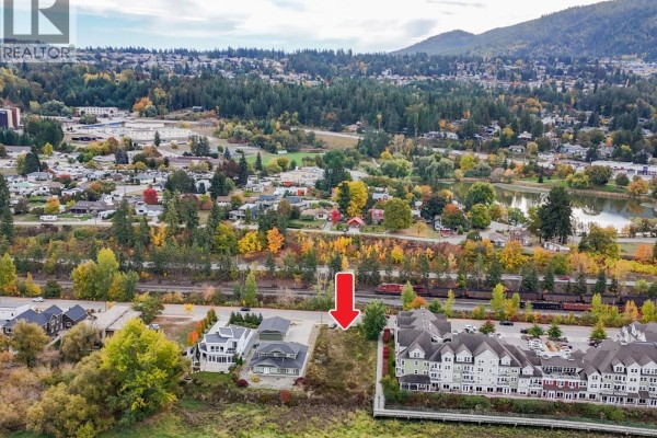 Aerial view of a vacant lot marked with a red arrow, with apartment buildings, houses, and forested hills in the background. - 721 Harbourfront Drive, Salmon Arm