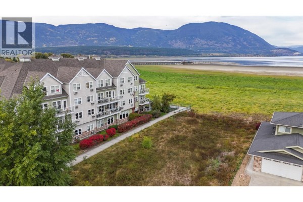 Aerial view of a vacant lot next to a beige apartment building, with a lake and distant mountains in the background. - 721 Harbourfront Drive, Salmon Arm