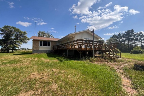 Rear view of a cream-colored house with a large wooden deck and stairs surrounded by green grass. - 72140 96 Rd N Road N, Plumas