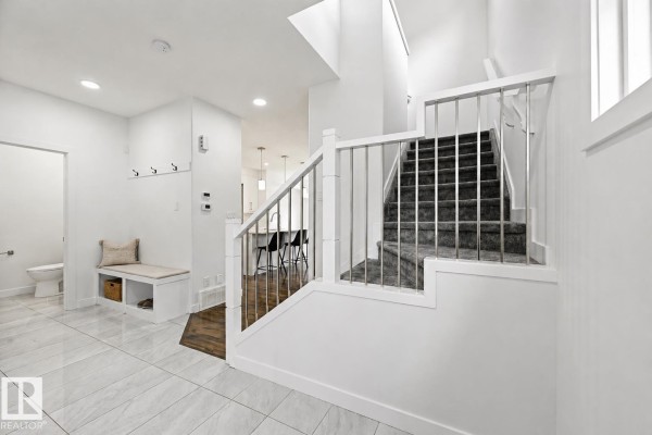 Bright entryway with grey carpeted stairs and a built-in bench. White tile floors extend to the kitchen. - 7258 Chivers Pl Sw, Edmonton