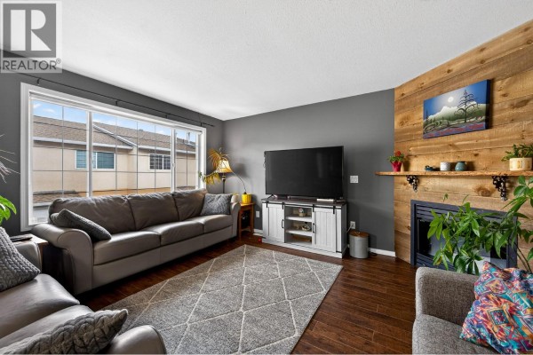 Living room with a gray sectional sofa, fireplace with wood accent wall, and a large window. - 726 Dunrobin Drive Unit# 1, Kamloops