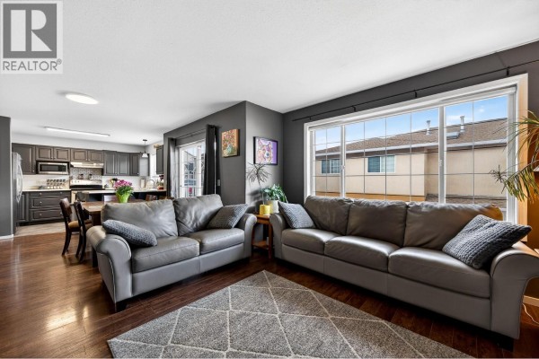 Living room and kitchen area with two gray sofas, dark wood floors, and a large window view. - 726 Dunrobin Drive Unit# 1, Kamloops