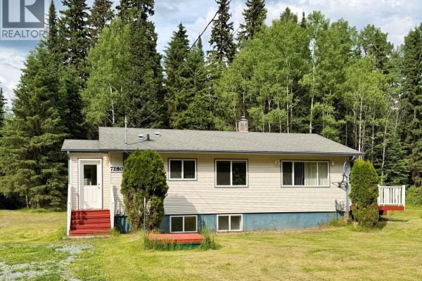 Side view of a beige bungalow with a green roof and a white deck, nestled amongst dense green trees. - 7280 Otway Road, Prince George