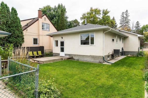 Back exterior view of a bungalow with a wooden deck, green grass, and a chain-link fence. - 729 Kildonan Drive, Winnipeg