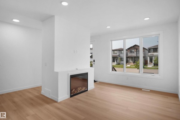Living room with a white fireplace, light wood floors, and a large window overlooking the street. - 73 Sienna Bv, Fort Saskatchewan