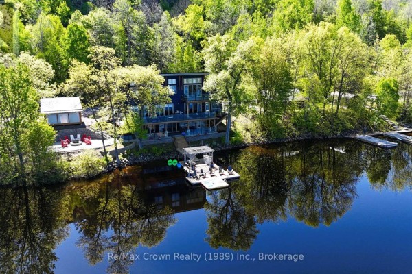 Expansive aerial view of the blue house and dock nestled in a wooded shoreline. - 7313 534 Highway, Parry Sound Remote Area