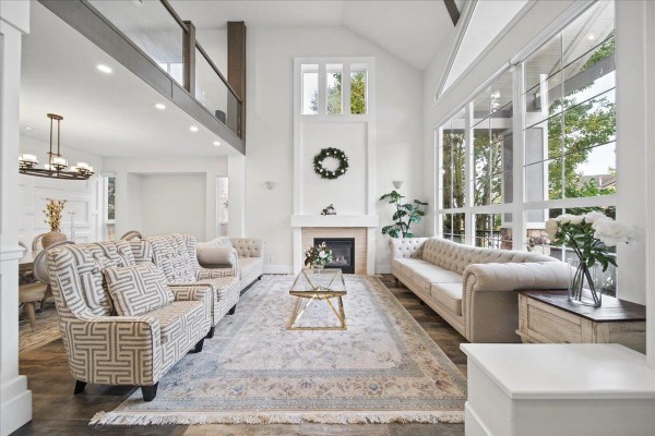 Living room with vaulted ceiling, fireplace, large windows, and two sofas with patterned armchairs. - 7355 146a Street, Surrey