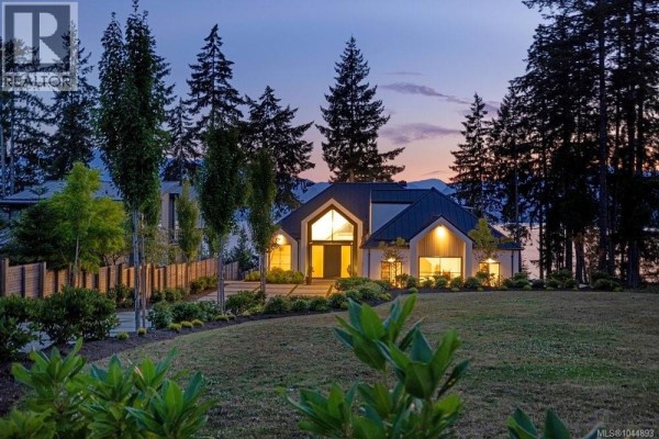 White modern home with peaked dark roof nestled amongst tall green trees and manicured lawn at dusk. - 7380 Lakefront Dr, Lake Cowichan