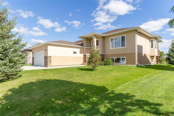 Side view of a beige stucco house with a two-car garage and manicured lawn. - 74 Breezy Bend, Steinbach