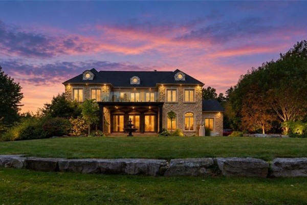 Exterior view of a large stone house with a balcony and fountain at dusk. - 740 Fiddlers Green Road, Ancaster