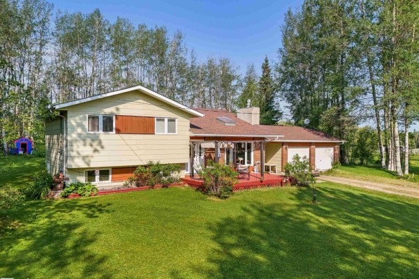 Side view of a yellow house with a red deck, red roof, and attached garage in a grassy, treed setting. - 7415 Twp Road 494 - House B, Rural Brazeau County