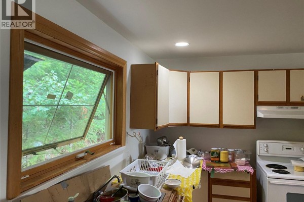 A kitchen with light beige cabinets, a wooden window frame, and a white stove overlooking a forest view. - 7475 Anglemont Way, Anglemont