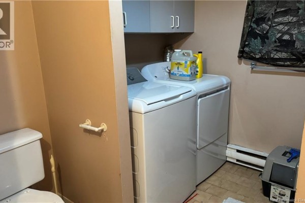 Laundry room with a white washer and dryer, tan walls, grey cabinets, and a white toilet. - 75-77 Belisle Street, Edmundston