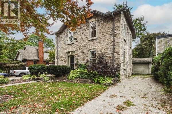 Side view of stone house with detached garage, driveway, and lush landscaping under a cloudy sky. - 75 Pollock Avenue, Cambridge